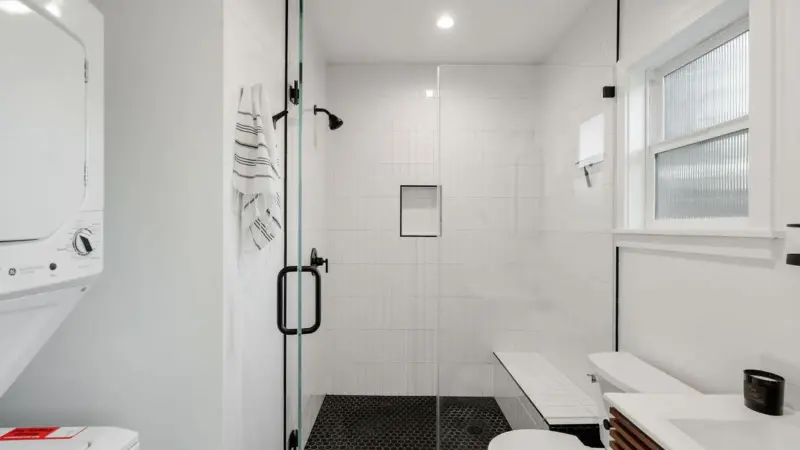 Burbank ADU bathroom with frameless glass walk-in shower, black hex tile floor, matte black fixtures, built-in bench, and stacked in-unit washer and dryer