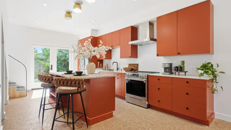 Culver City ADU kitchen with custom terracotta flat-panel cabinetry, black hardware, brass-toned ceiling lights, a speckled island top, and cork floors