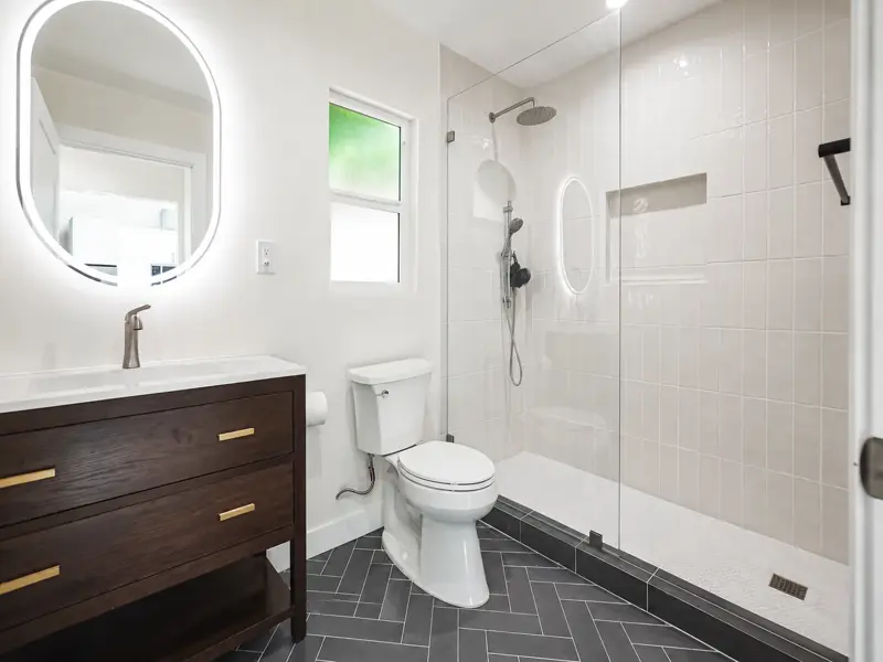 Designer bathroom in the Mar Vista ADU with a walnut vanity, backlit oval mirror, charcoal herringbone floor tile, and a frameless walk-in shower