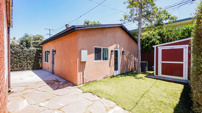 Side yard of the Mar Vista ADU with flagstone walkway, artificial turf, garden shed, and the terracotta stucco elevation