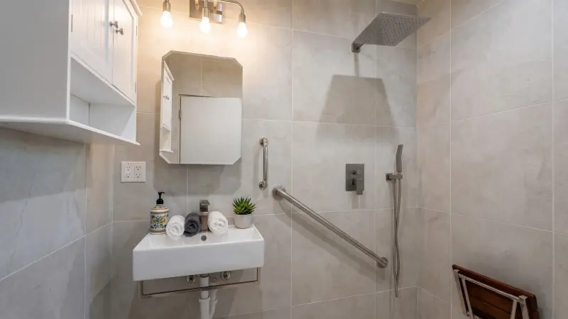 Accessible bathroom in the West LA HPOZ Wilshire ADU with a curbless roll-in shower, stainless grab bars, fold-down teak bench, and a wall-hung sink with knee clearance