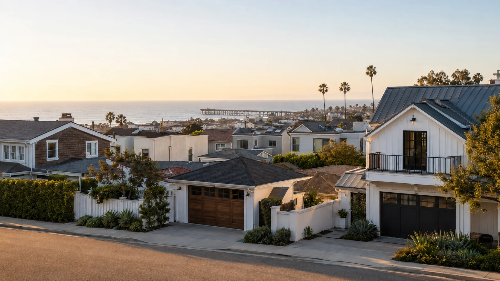 Modern white coastal homes on a Hermosa Beach residential street at golden hour, with mature palms and the Hermosa Beach Pier silhouetted at the Pacific horizon