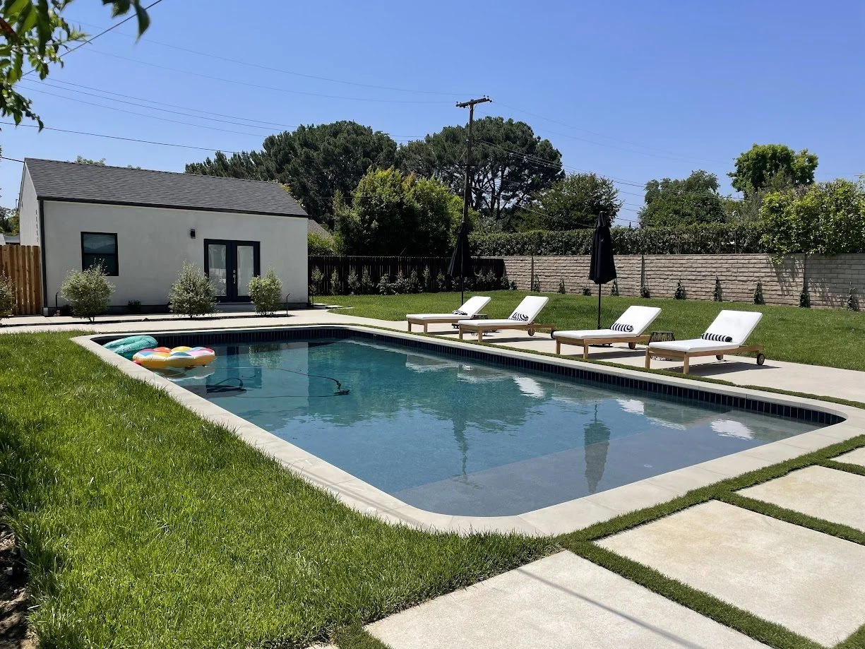 White stucco gable pool house ADU with French doors and a lineup of chaise lounges across the pool deck